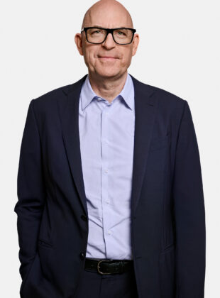 A man in a dark suit and light blue shirt against a white background.