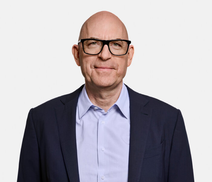 A man in a dark suit and light blue shirt against a white background.