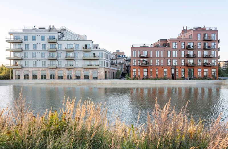 Modern apartment buildings beside water with greenery in the foreground.