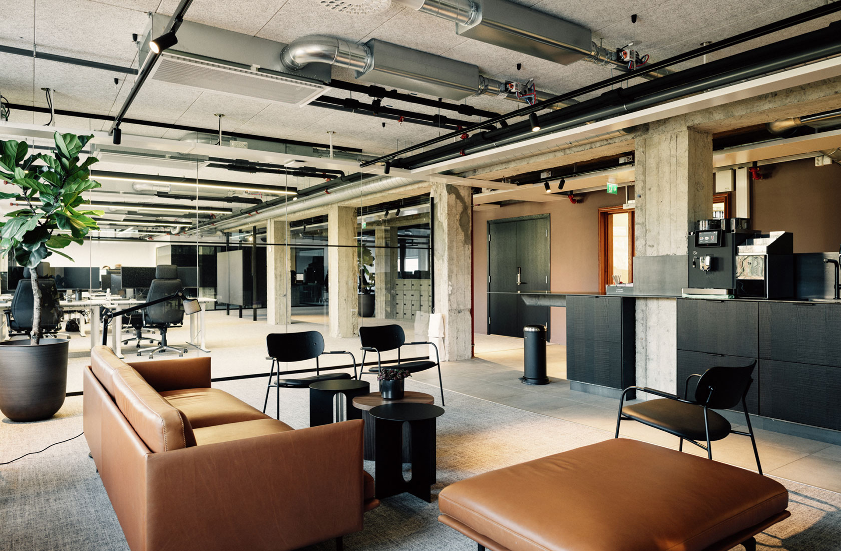 Office space showing a lounge area with sofa and chairs, pillars of concrete and a conference room in the background.