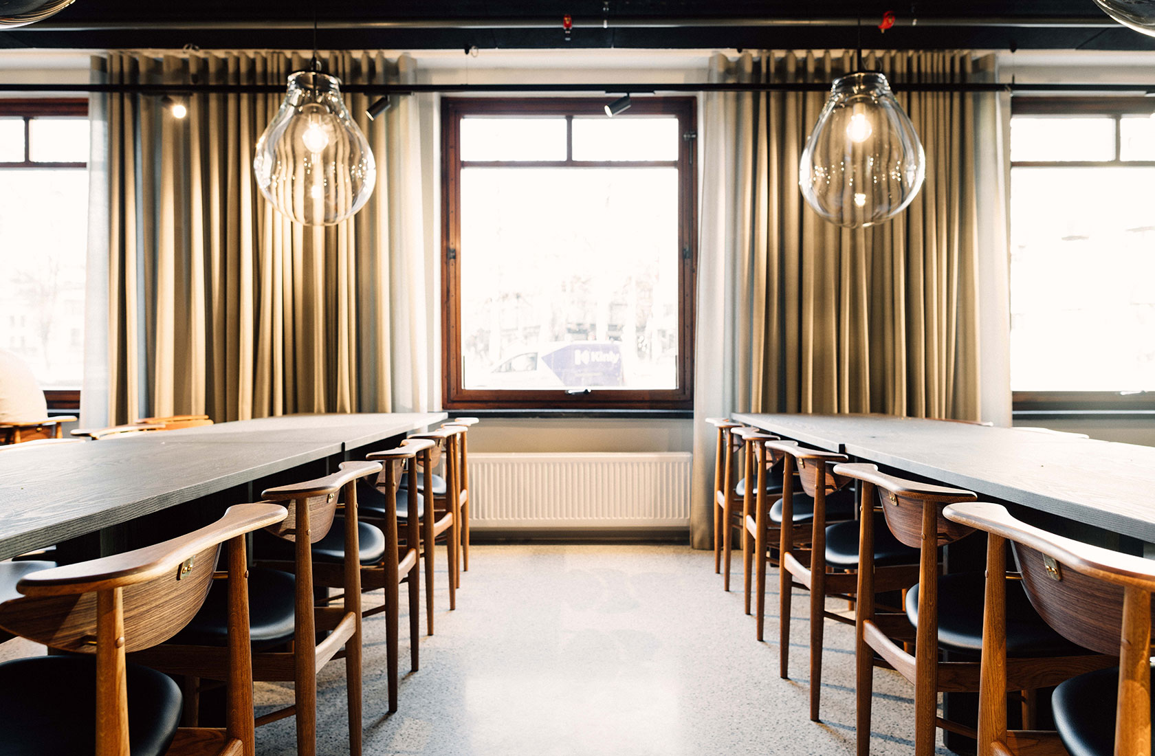 Two long conference tables with a lot of charis made of wood. Windows in the background and long beige curtains between the windows.