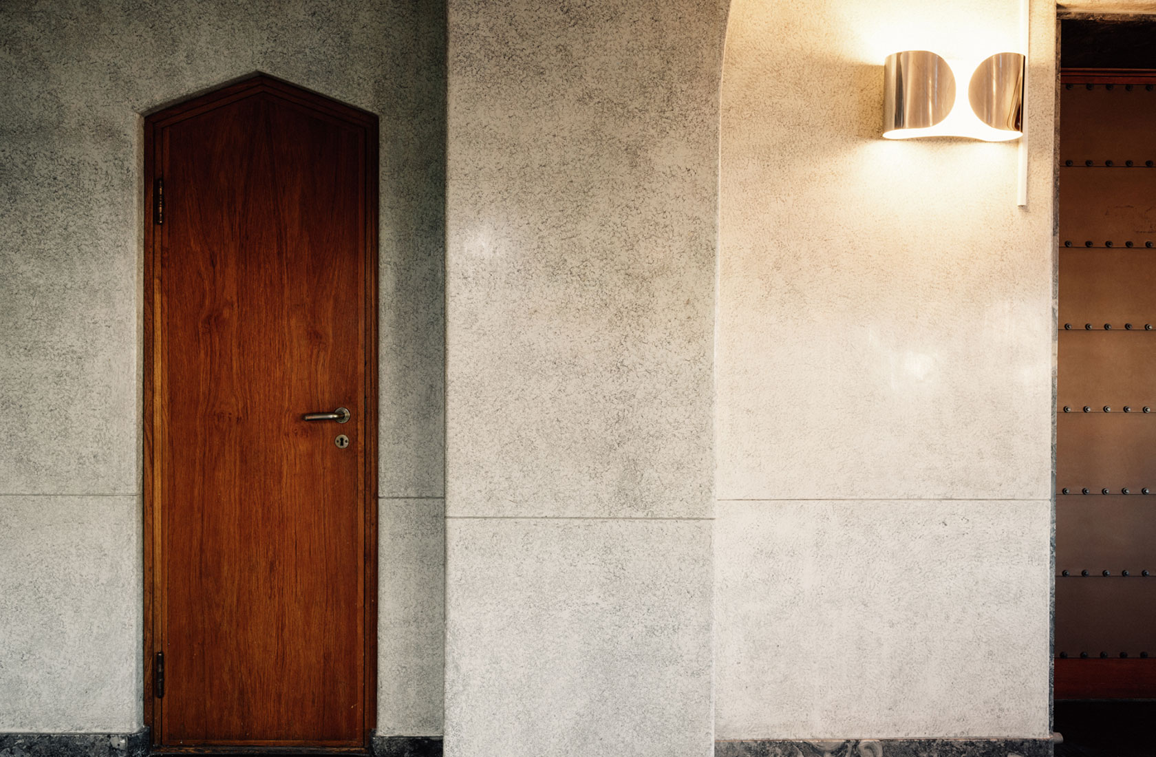 Close up of a wall made of stone with a wooden elderly door on the left side and a lamp placed on the wall on the right side.
