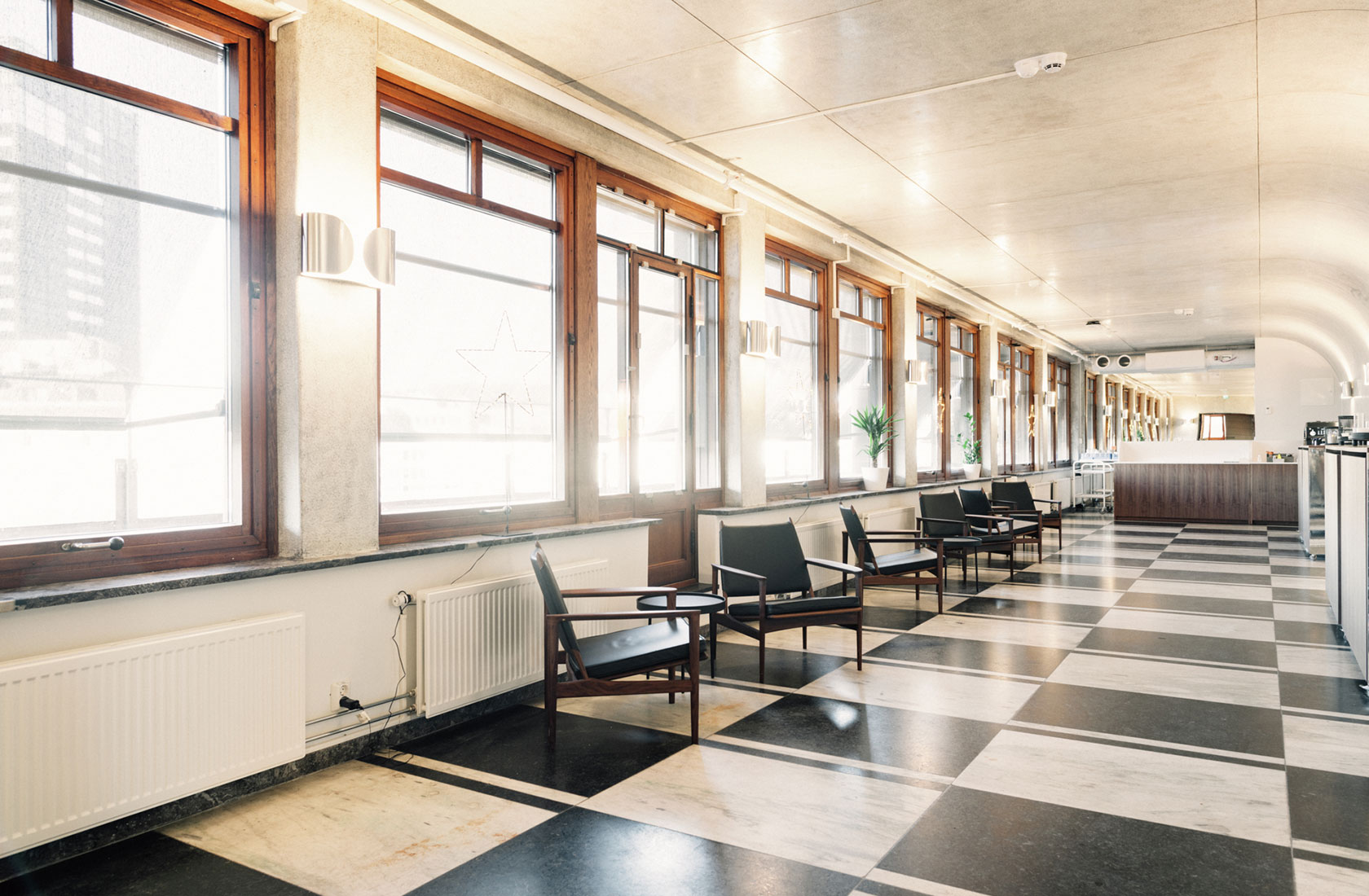 Office space showing an open space with a floor made of squares in black and white stone. Big windows and black chairs in groups.