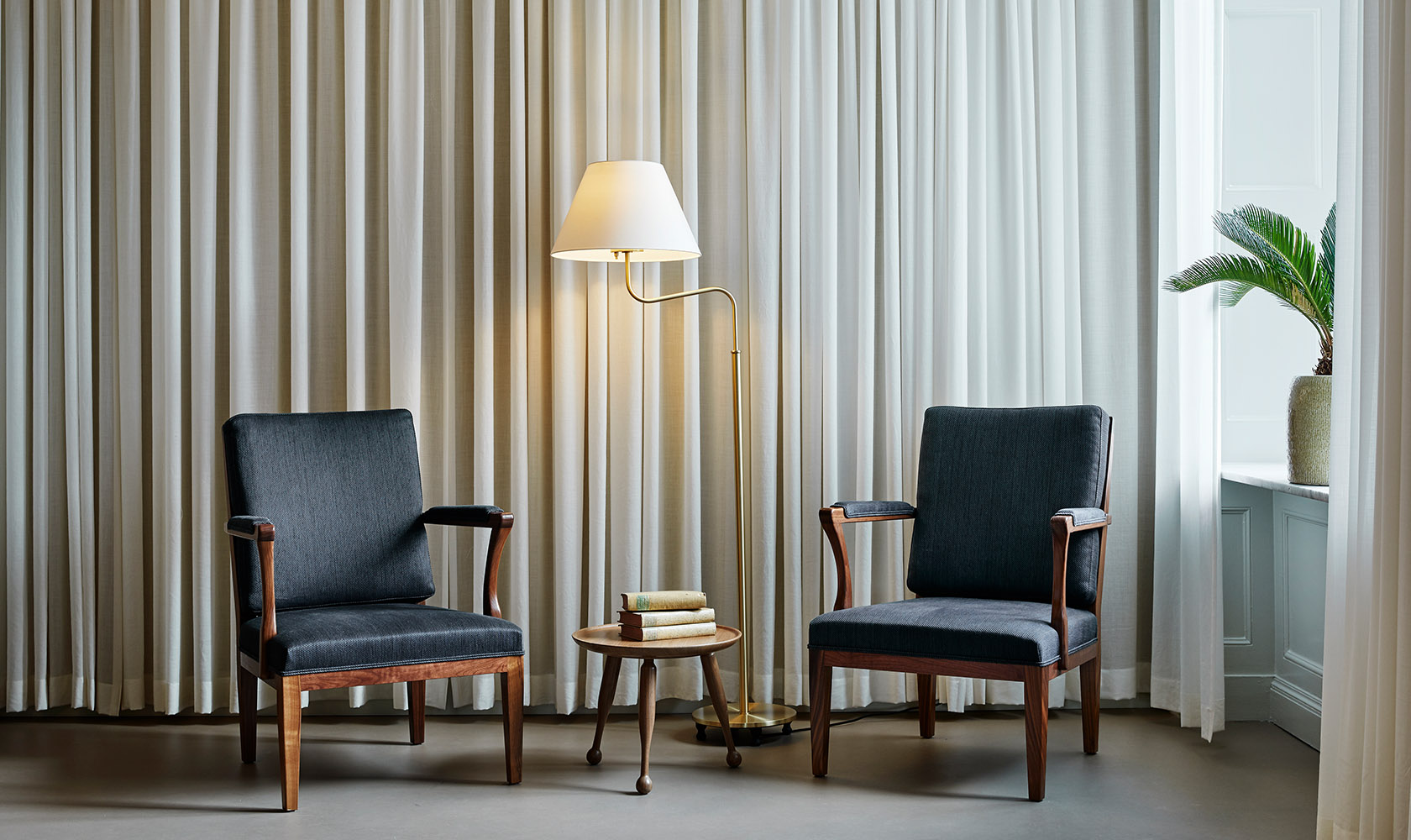 Two armchairs and a small table in the middle and a floor lamp in front of a fabric in off-white. To the right a window with a green plant.