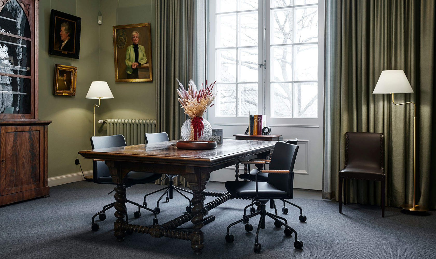 An older wooden conference table with four modern chairs on wheels. In the background an older, larger window with white details. On the sides of it curtains in green. In the left corner, older paintings and an older cabinet in darker wood with glass details.