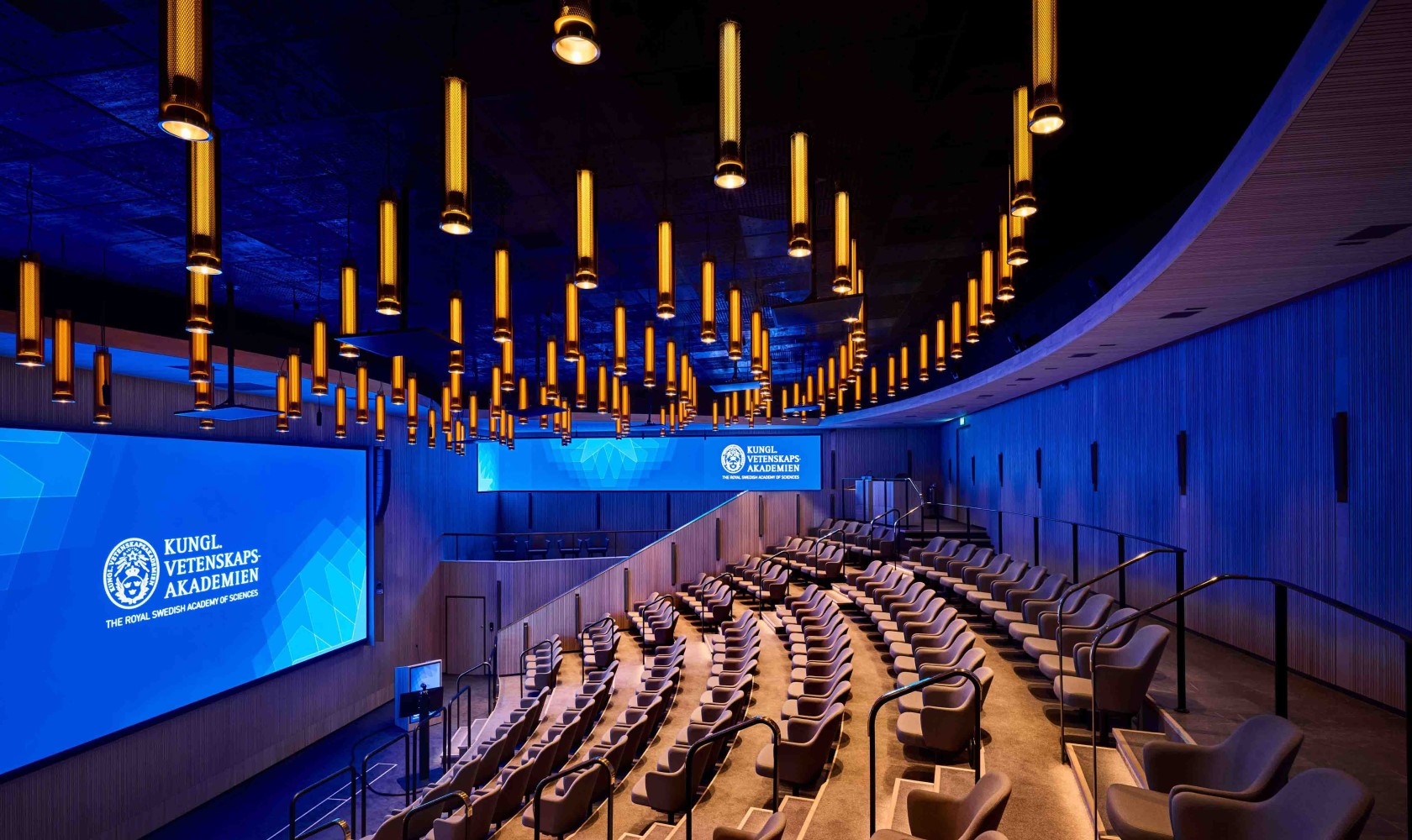 Auditorium with modern chairs in beige. Larger led screen shows in front, a smaller stage and a smaller leed scrren one on the short side. Interior in light wood. A large number of light sources in the ceiling that resemble golden rods.