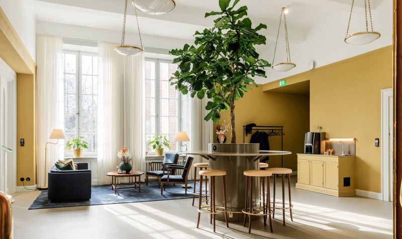 Lounge room in older style with ocher colored walls and ceiling and wooden details in white. At the far end two large windows and in front a sofa, table and two armchairs. To the right, a coffee station in ocher and in the foreground a round bar table with a larger plant in the middle and a number of bar stools.