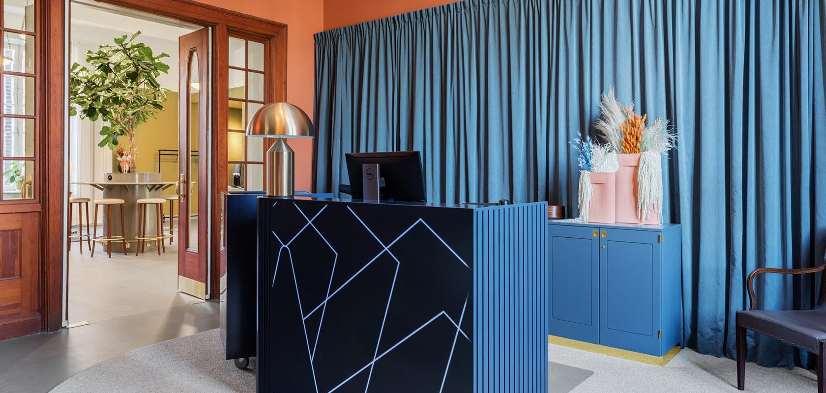 Reception area with reception desk in blue. Brick-colored walls with a blue fabric behind the counter and cabinets in blue. A chair to the right and a door in a glazed part to the next room.