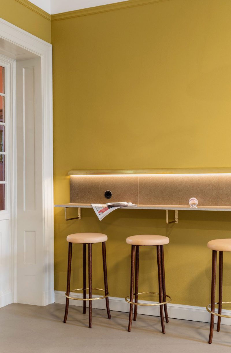 Ocher colored wall with a longer wall-mounted table with three barstools in front. Older door frame on the left in white.