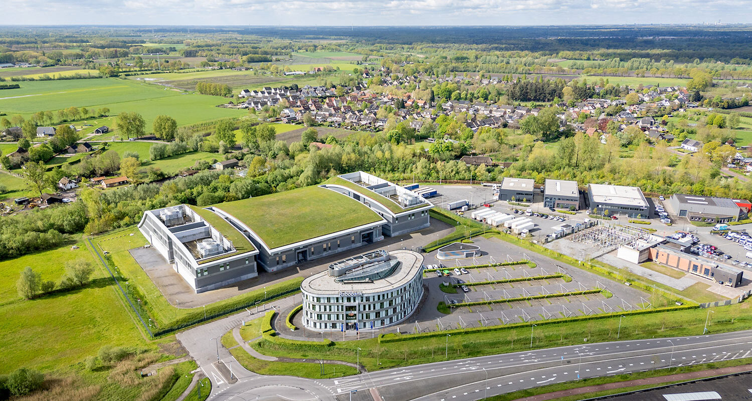 Data center located in a large single-story building with a white facade and a green roof. In front of it is an organic-shaped four-story office building with a larger parking lot to the right. There is a multi-lane road in front of the building with a roundabout to the left.