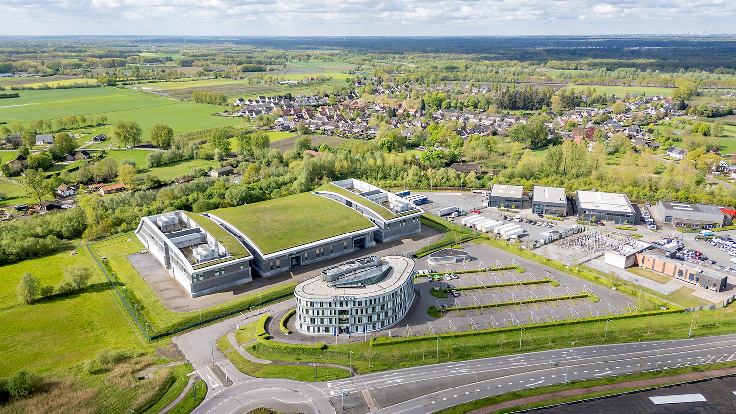 Data center located in a large single-story building with a white facade and a green roof. In front of it is an organic-shaped four-story office building with a larger parking lot to the right. There is a multi-lane road in front of the building with a roundabout to the left.