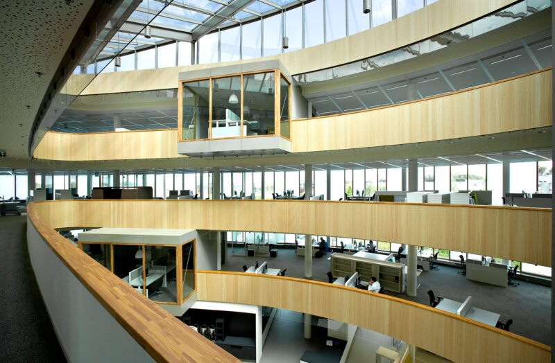 View from the multi-story office building that has an open ceiling in the middle. The surfaces of the floors facing the central area are made of light wood, giving the building its character. There are large windows along the facade.