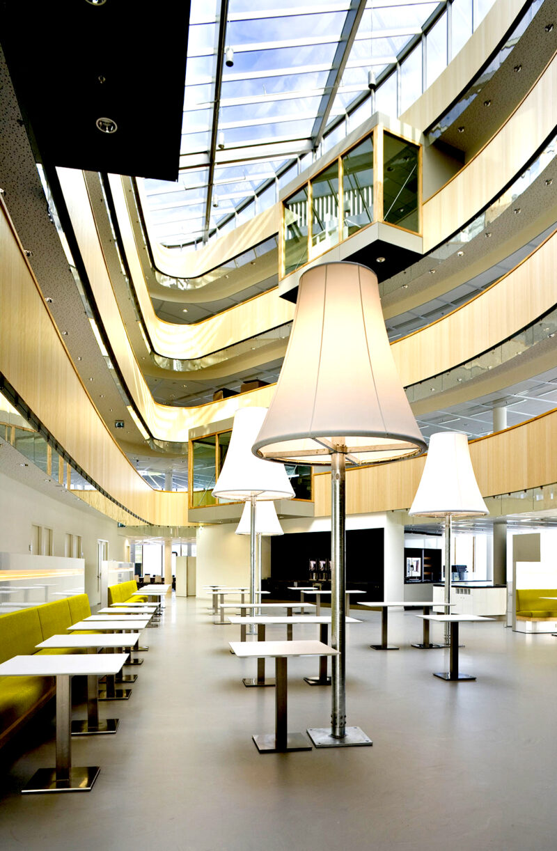 View from the ground floor of the office building with green sofas and several tables. Several floor lamps with large white shades are scattered in the middle. You can see up towards the floors from the open central area and the light wood sides that mark the floors.