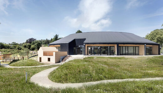 A kindergarten on a hill in a circular building made of light wood with a black roof and black facade sections surrounded by grass.