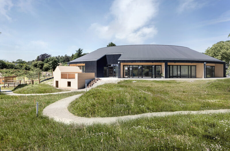 A kindergarten on a hill in a circular building made of light wood with a black roof and black facade sections surrounded by grass.