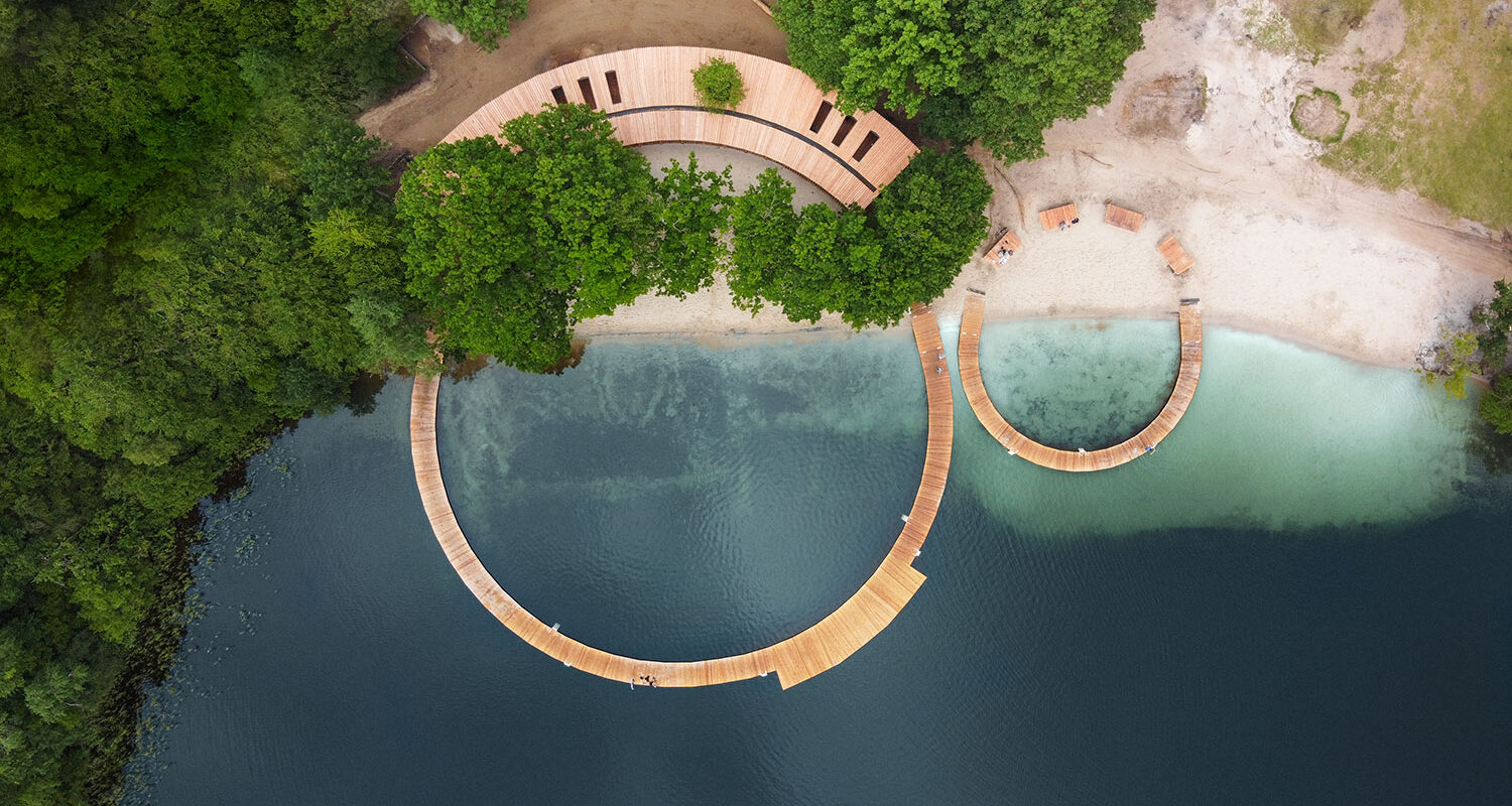 View from above of wooden docks forming two circles of different sizes. In one part of the larger circle, the docks transition into a building with a roof. Beautifully located by calm water and lush forest.