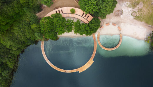 View from above of wooden docks forming two circles of different sizes. In one part of the larger circle, the docks transition into a building with a roof. Beautifully located by calm water and lush forest.