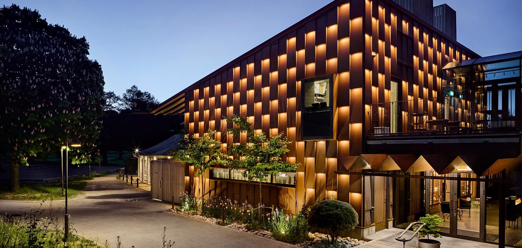 Evening photo of illuminated facade of a restaurant in rust-colored corten steel. The building is two stories high with a sloping roof.