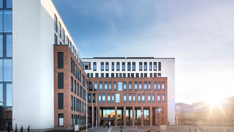 Exterior image of a hospital building with six floors, white plaster, and a protruding section in red brick against a clear blue sky. A large part of the facade is covered with vertical windows.