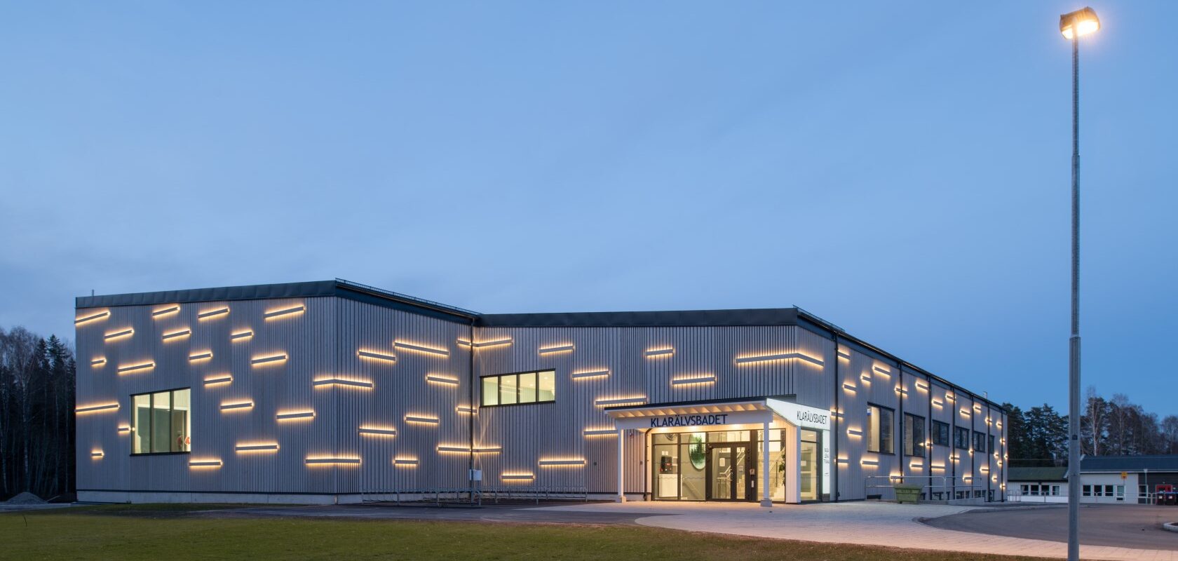 Evening picture of a swimming hall with vertical wooden panels of different widths forming a pattern. Details with horizontal elements of illuminated wooden panels. Welcoming entrance with large glass sections, trimmed lawn around the building.