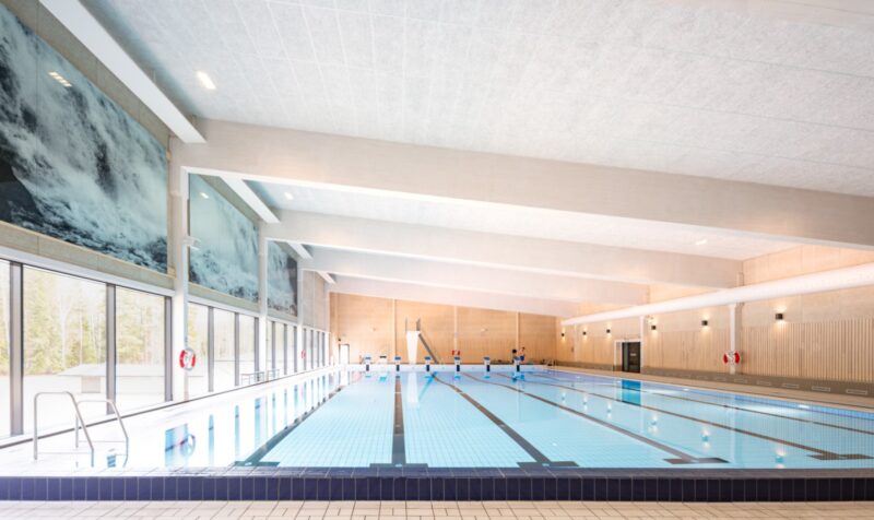 Interior image of a swimming hall with a six-lane pool and a diving tower. Walls in lighter wood paneling and white ceiling.