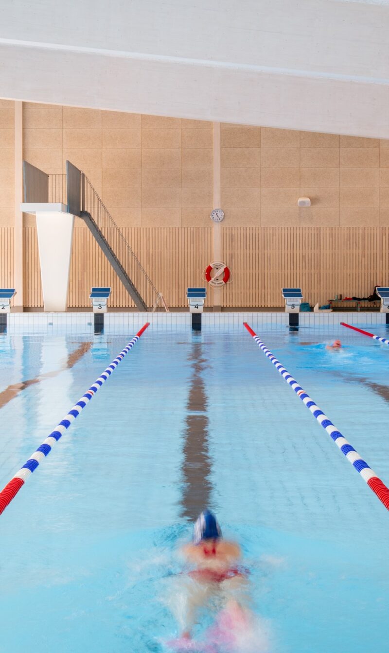 Interior image of a swimming hall with a person swimming, diving tower in the background. Walls in lighter wood paneling and white ceiling.