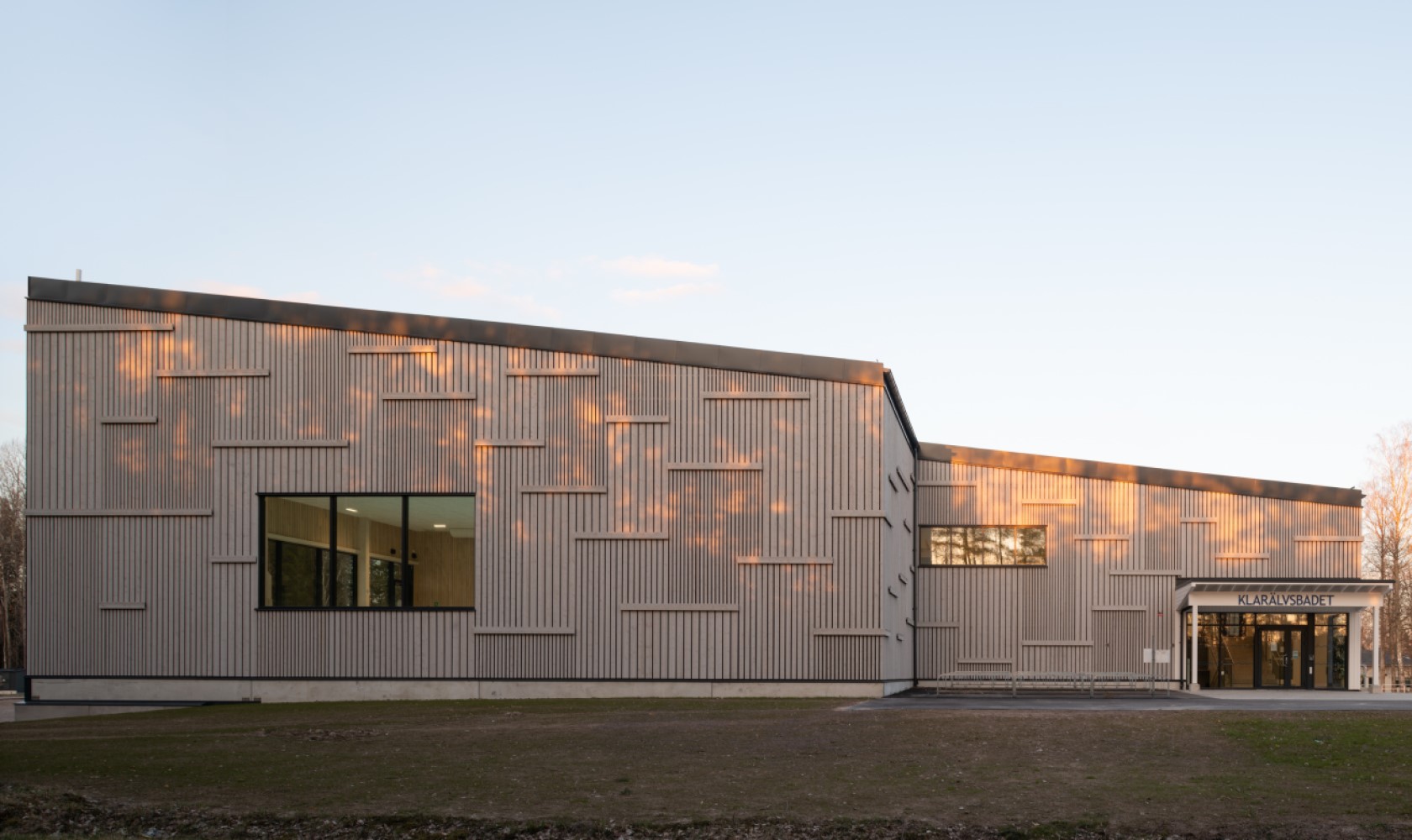 Swimming hall with vertical wooden panels of different widths forming a pattern. Details with horizontal elements of wooden panels. Entrance on the right with glass sections.