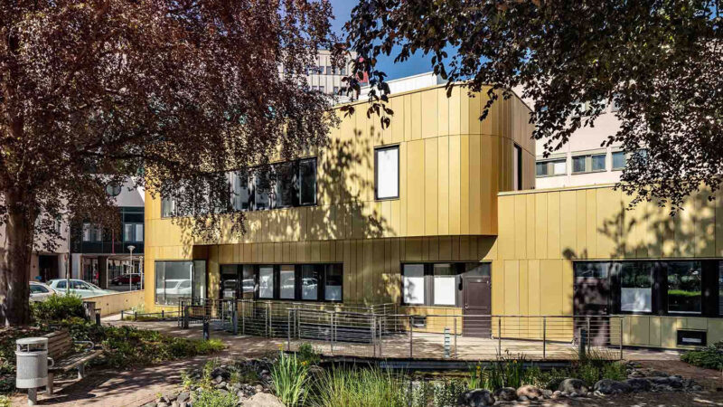 Organically shaped two-story building with a facade in yellow-colored metal. Park-like area in front of the building with vegetation and beautiful stone elements.