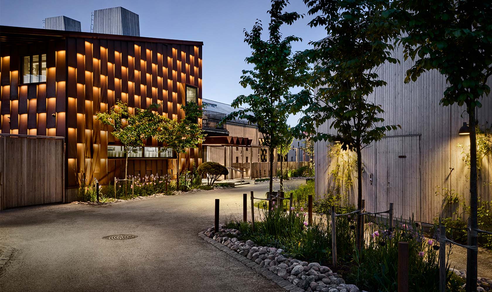 Evening photo where the facade and surroundings are nicely illuminated, restaurant building in the background with a facade in corten steel.