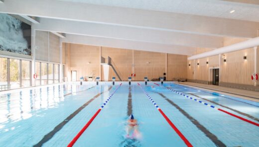 Interior image of a swimming hall with a pool in which a person is swimming. The pool has six lanes and a diving tower at the far short side. Walls in lighter wood paneling to the right and large glass areas to the left.