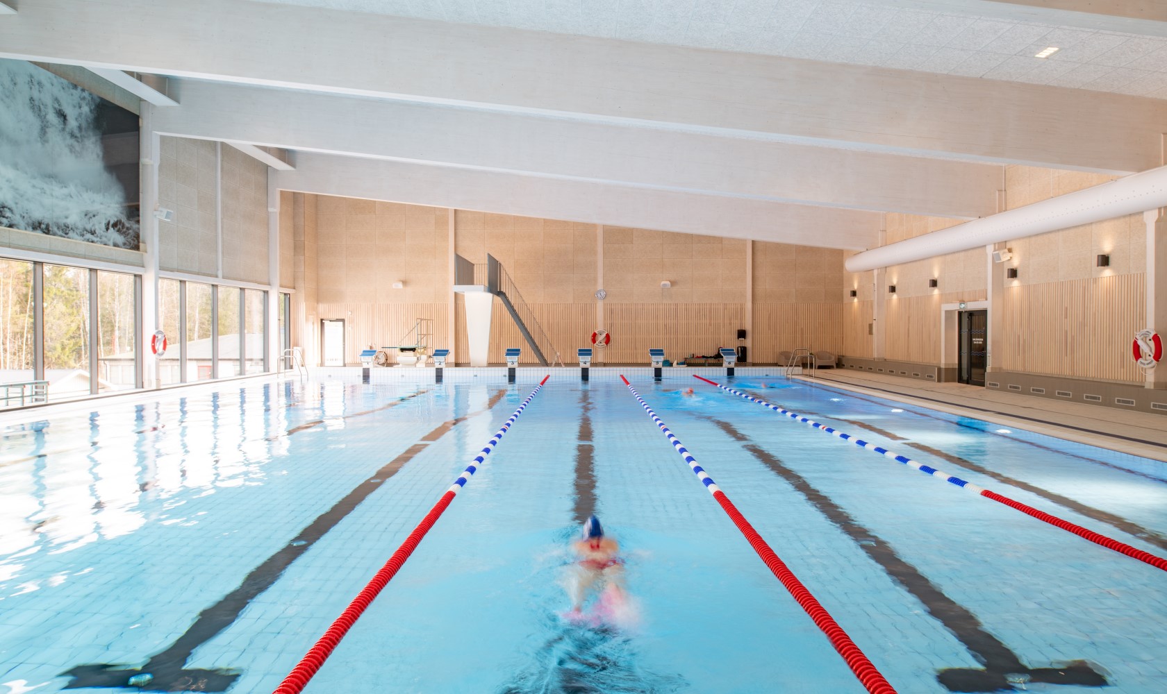 Interior image of a swimming hall with a pool in which a person is swimming. The pool has six lanes and a diving tower at the far short side. Walls in lighter wood paneling to the right and large glass areas to the left.