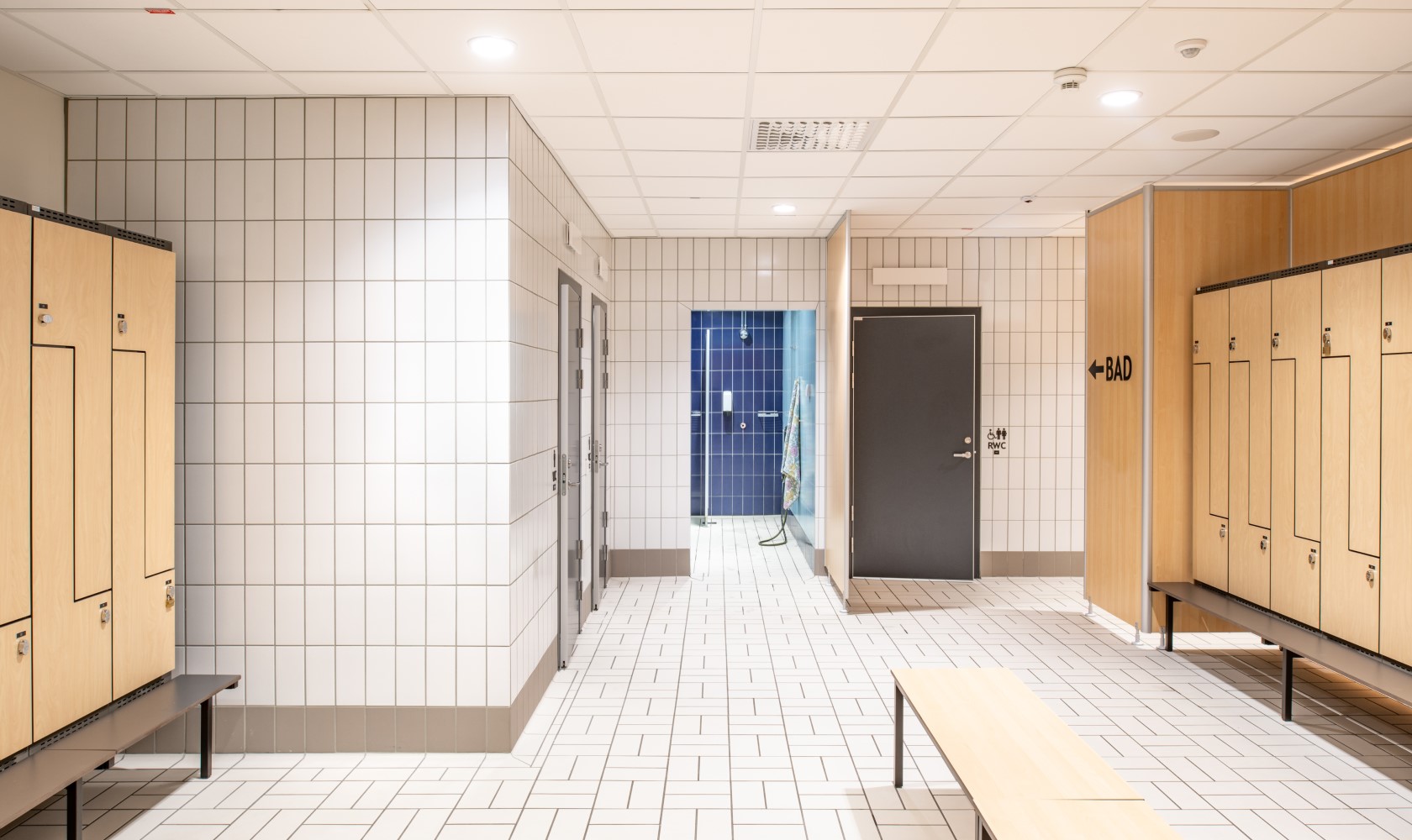 Changing room with white tiled walls and white tiled floor, light-colored wooden lockers. Shower area in blue can be seen in the background.