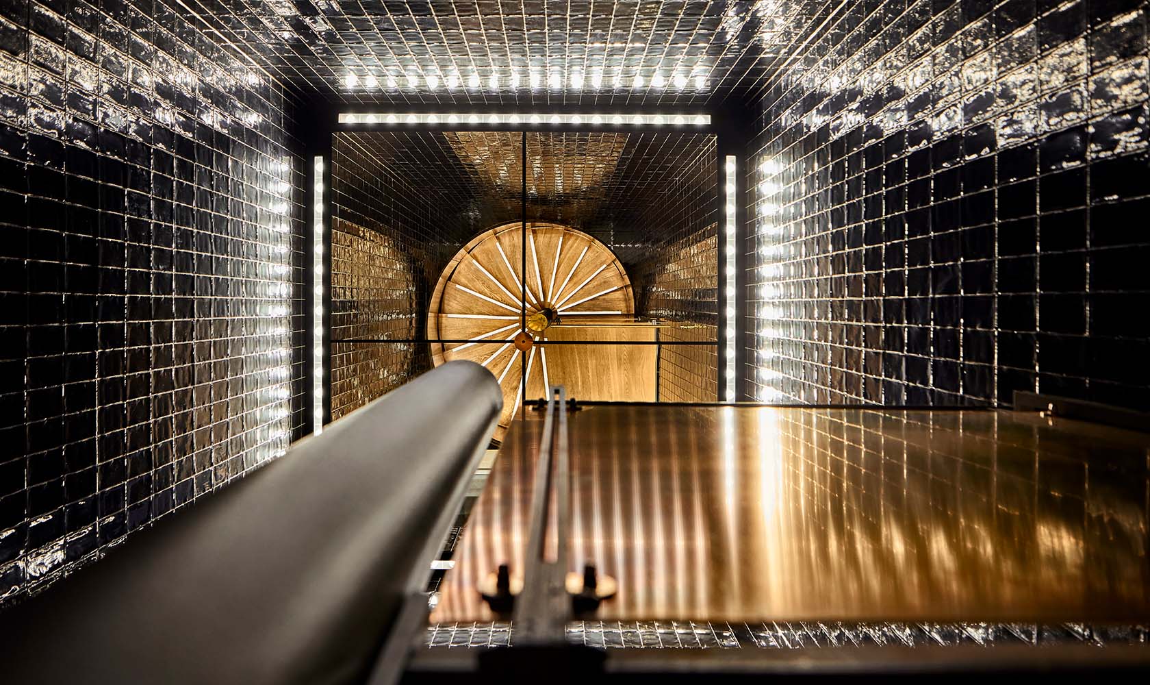 Beautiful lighting design in a narrow staircase photographed from the bottom where the mirror ceiling reflects the oak staircase. Walls of black tiles.