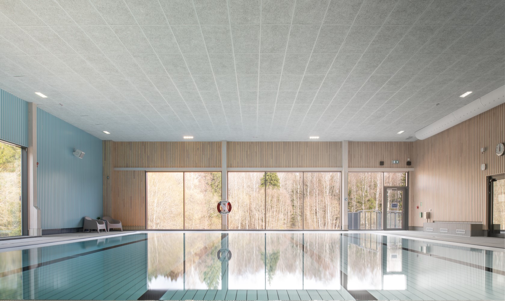 Interior image of a swimming pool with calm water. Walls in lighter wood, the far wall has a larger glass section facing a forest.