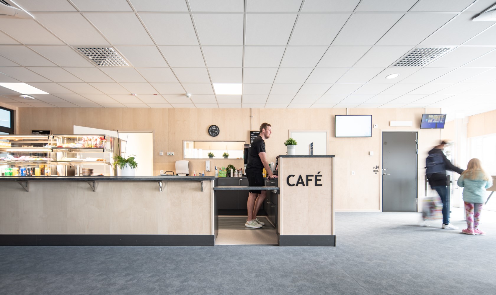 Café picture from the swimming hall facility. Counter and walls in lighter wood with a gray floor. One person behind the counter and two people walking beside it.