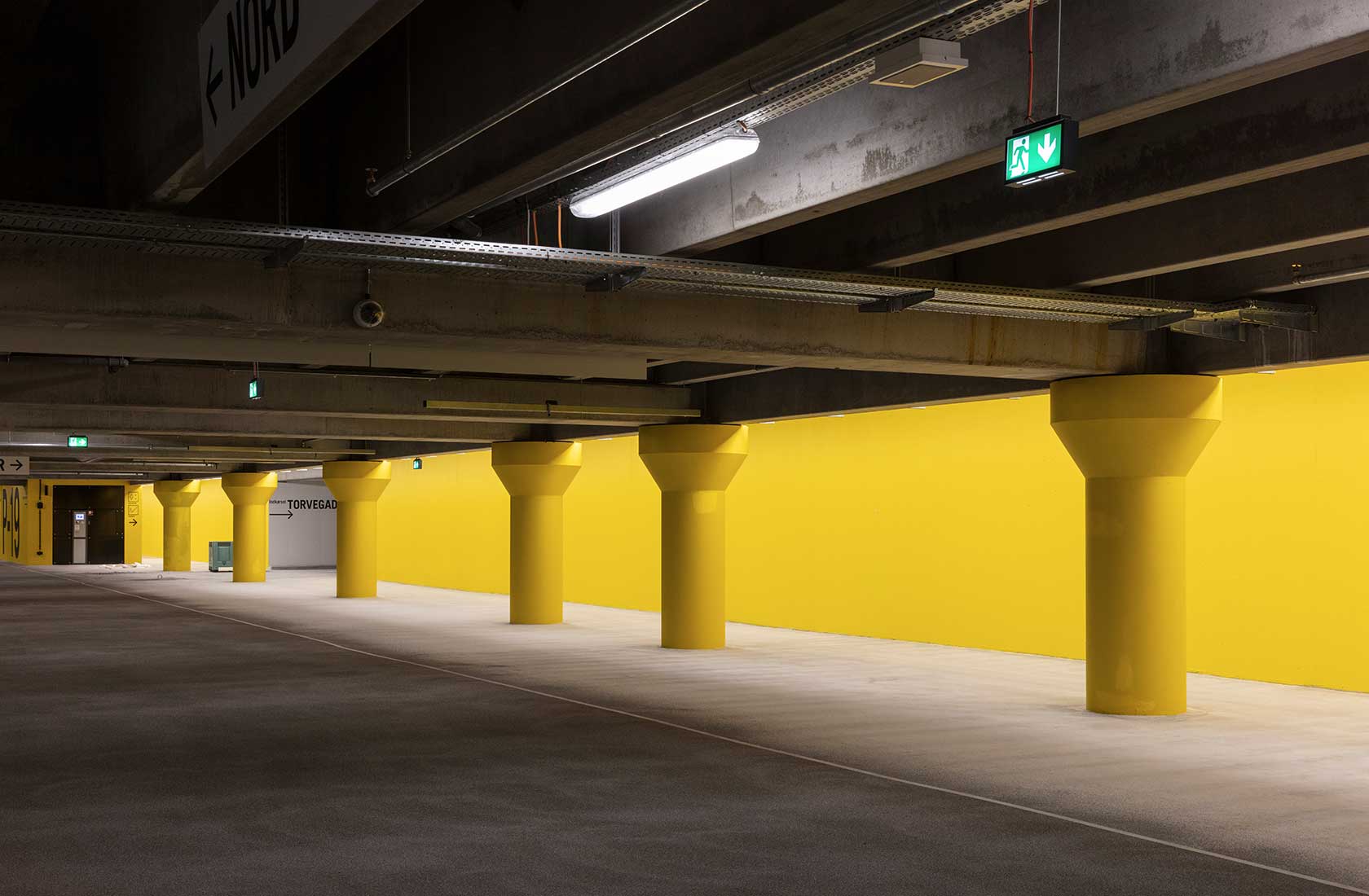 A parking garage with several pillars and a yellow wall behind it.