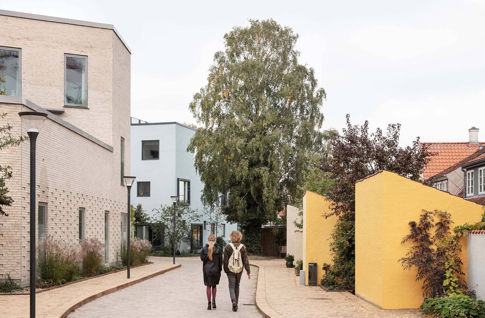 Two women walking on a pedestrian path, to the left a lighter brick house with three stories. Buildings around in different colors and shapes.