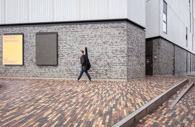 A man dressed in black with a guitar case on his back passes by a gray brick wall.