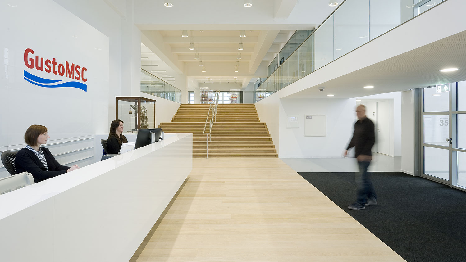 Reception area and entrance for the company GustoMSC, airy and bright with white walls and a sign behind the reception desk where two people sit. Stairs in light wood in the background.
