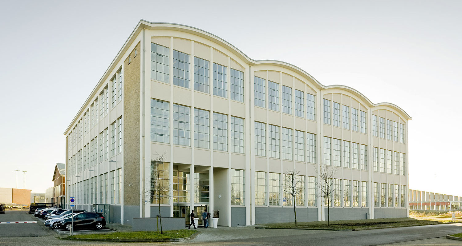 Renovated older industrial building in light yellow with a shell roof and large windows. Road ahead and parking along the left side.