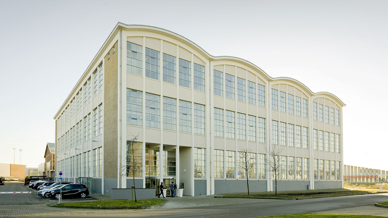 Renovated older industrial building in light yellow with a shell roof and large windows. Road ahead and parking along the left side.