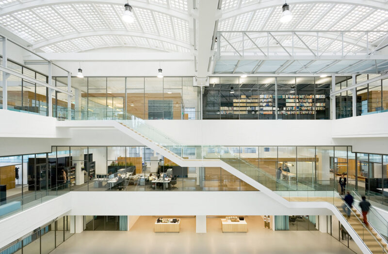 Interior image of an office building with a large atrium in the middle with a glass roof in the form of arched arches. A staircase that extends from the lower level up to level three. Airy office spaces in white, light wood and glass surfaces.