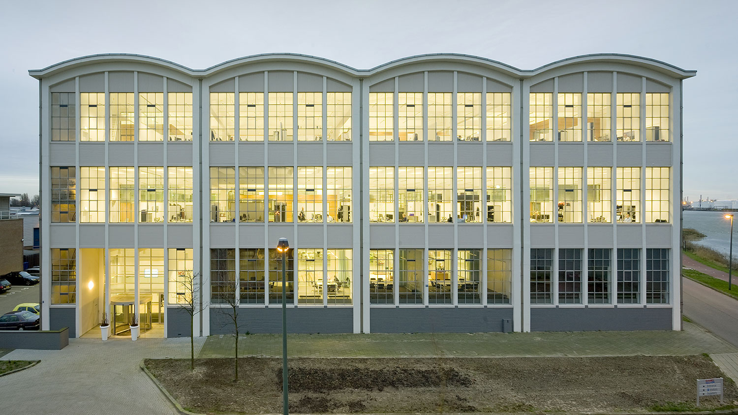 Evening picture from a renovated older industrial building in light yellow with a shell roof and large window sections, the sea can be seen on the right and parking on the left. Inside the windows you can see a modern office landscape.