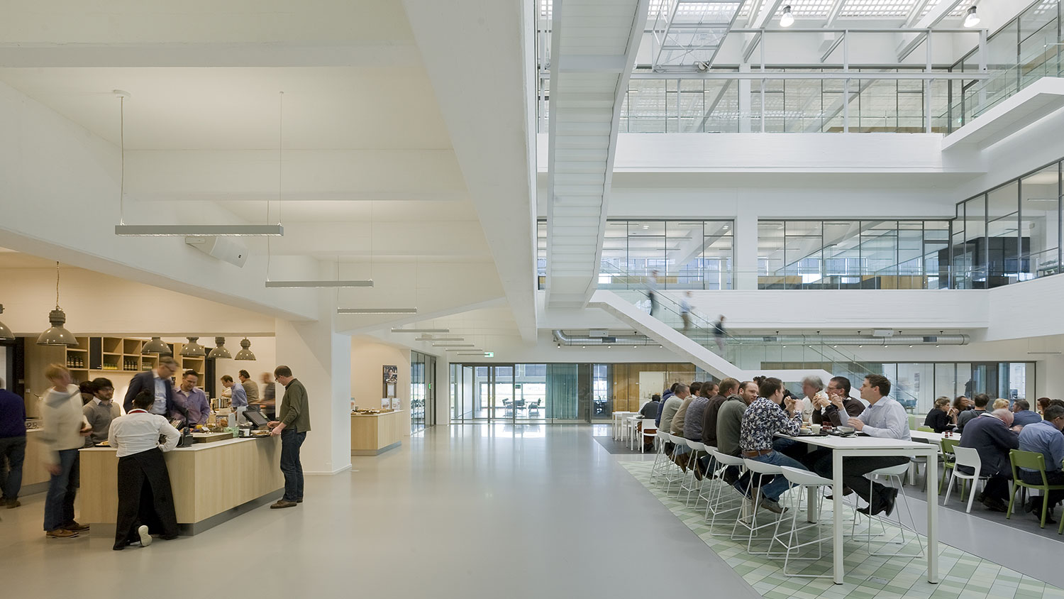 People serving themselves at buffet tables on the left and long tables with many people eating on the right at the ground floor of a modern office.