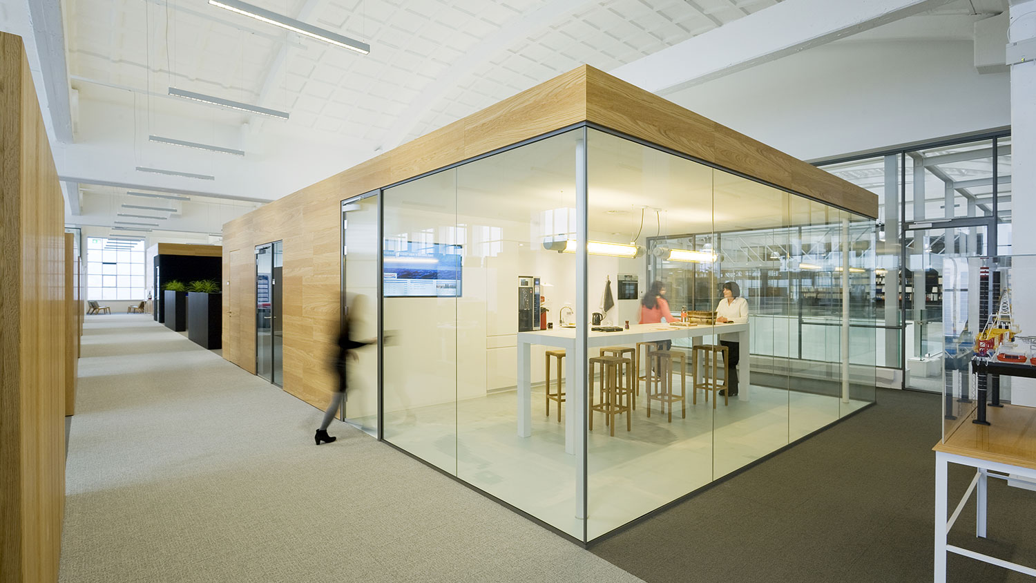 Corridors in an open modern office landscape, dividers in light wood on the left towards the workplaces. On the right, a person entering a small conference room with glass walls.