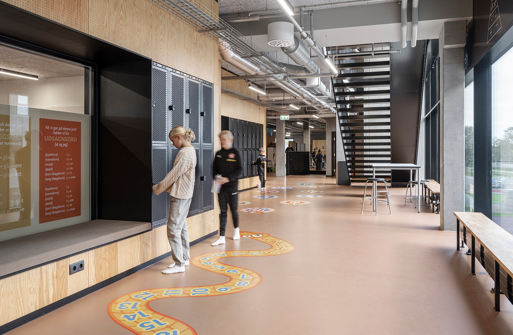 Interior image of the school building on the lower floor with a couple of children standing by a wall with black lockers and a black metal staircase leading up to the upper floor.