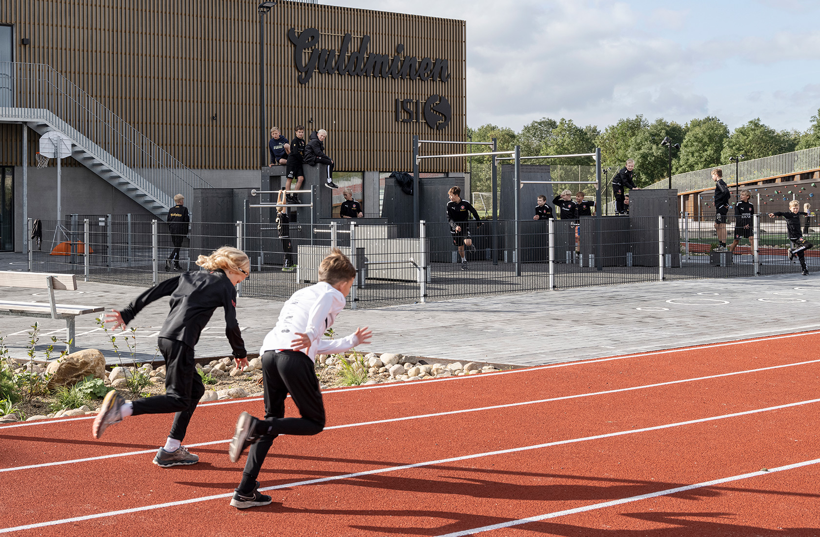 Two children running fast on a red running track with a school building in the background.