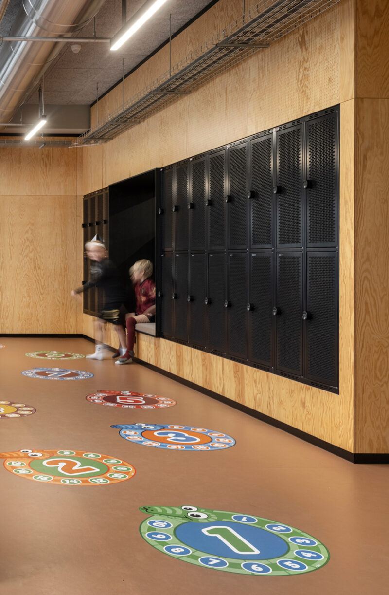 Interior image of a school building with black metal lockers on the far right side, plywood walls, and two children sitting in a space between the lockers.
