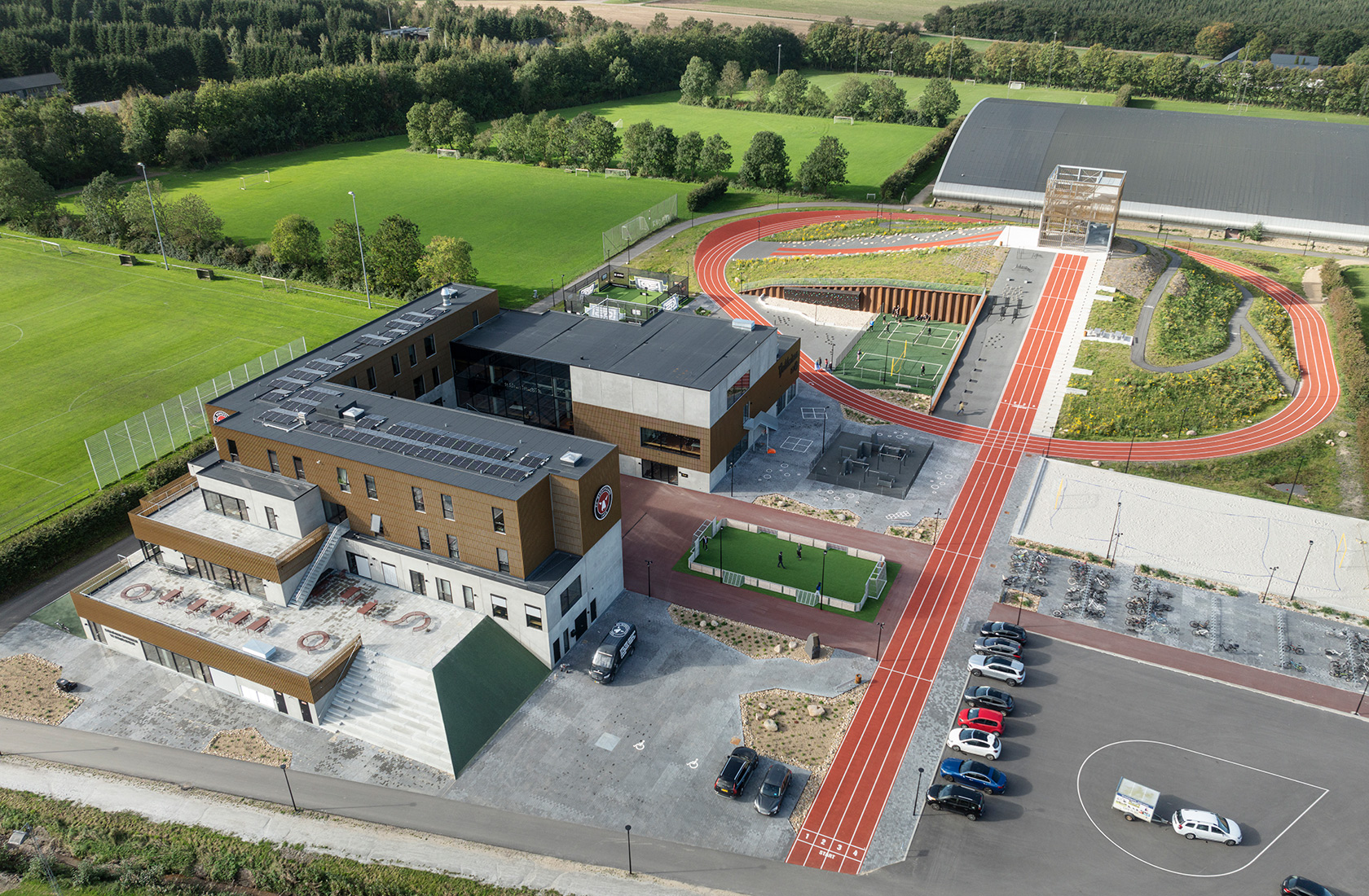 View from above of a sports facility with a larger property, red running tracks, playing fields, and other activity-based equipment, as well as a large parking lot.