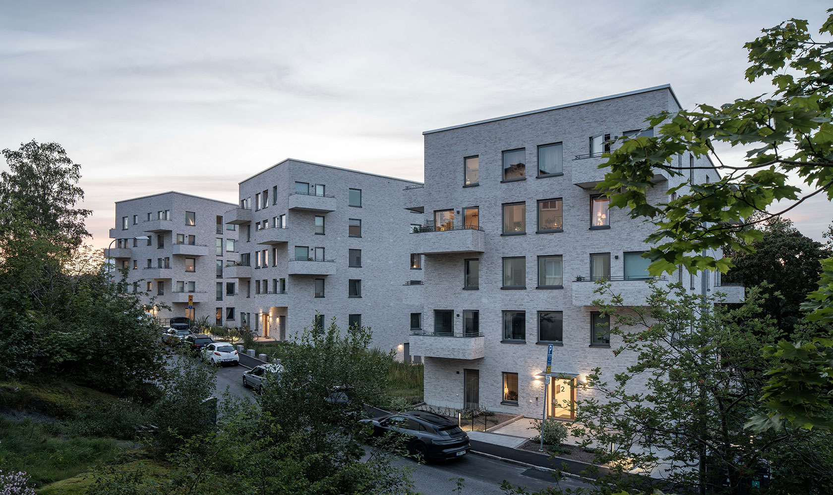 Evening shot of three five-storey buildings in beige brick, with green trees in the foreground.